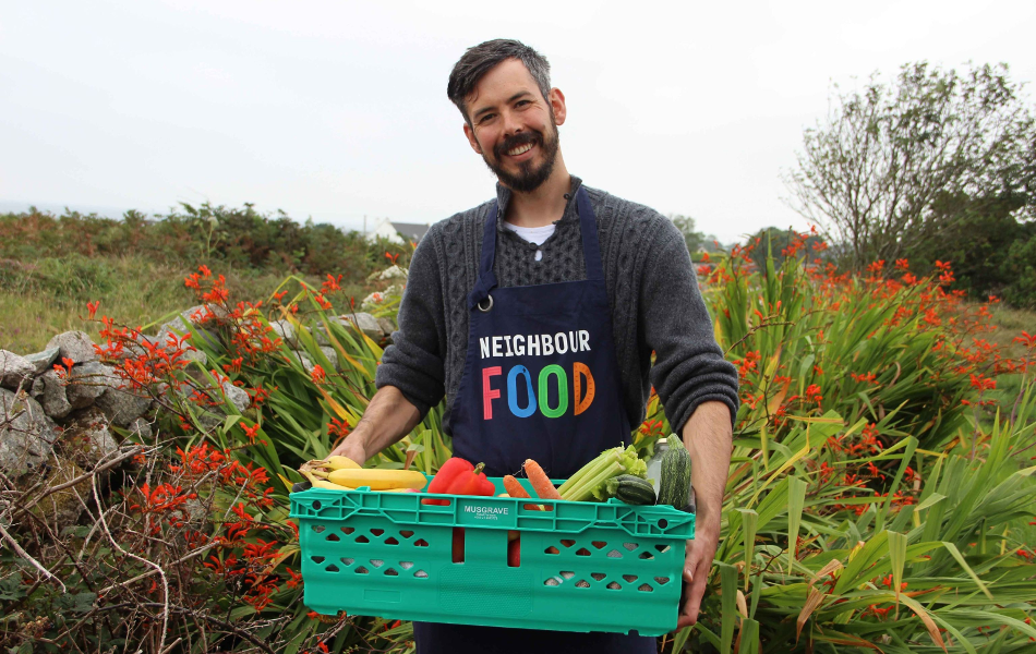 Diarmuid Ó Mathúna outisde between flowers holding a green box full of fruit and vegetables